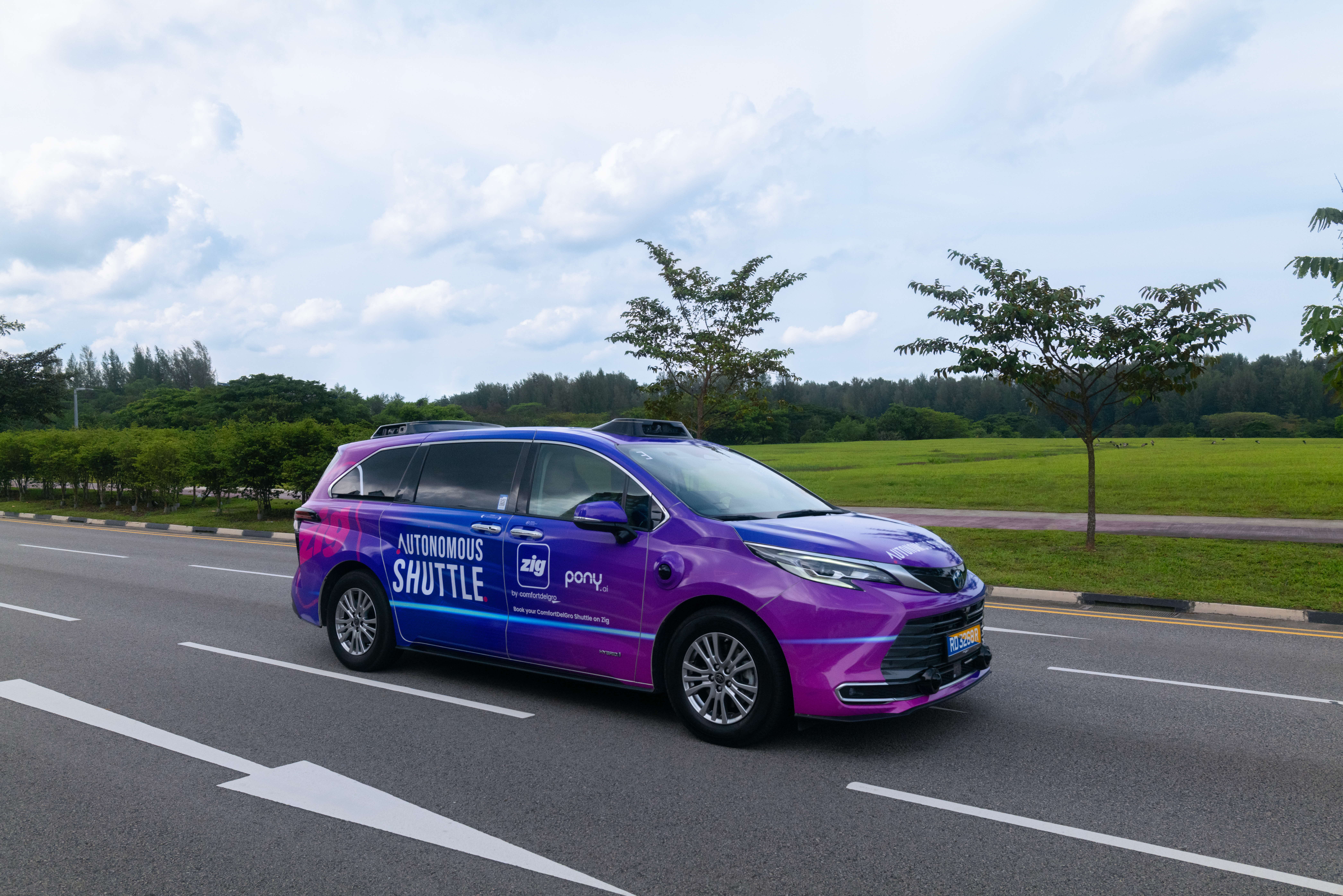 A blue-purple autonomous shuttle branded "ComfortDelGro Autonomous Shuttle" drives along an open road flanked by greenery.